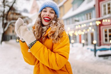 A happy woman with red lipstick has fun with the snow while walking along the snow-covered city streets. A beautiful tourist enjoys a sunny winter day. Concept: beauty, weekend, and lifestyle.