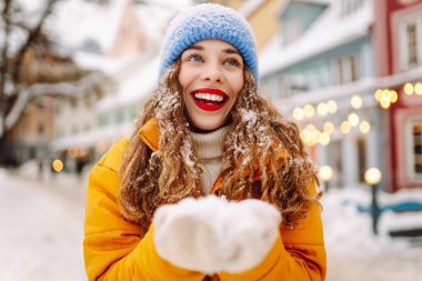 A happy woman with red lipstick has fun with the snow while walking along the snow-covered city streets. A beautiful tourist enjoys a sunny winter day. Concept: beauty, weekend, and lifestyle.
