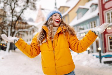 A happy woman with red lipstick has fun with the snow while walking along the snow-covered city streets. A beautiful tourist enjoys a sunny winter day. Concept: beauty, weekend, and lifestyle.