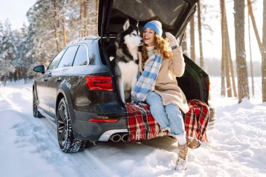A cheerful woman sits in trunk of a car in snowy forest with her pet. A woman and her Siberian husky are on a road trip on a sunny winter day. Concepts include adventure, relaxation, and weekends.