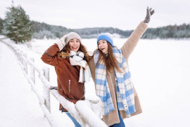 Two young women in warm clothes are having fun in a snowy forest by a lake. Friends are enjoying a sunny day, spending time together. Concept of fun and friendship. Winter day.
