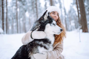 A young woman in a warm coat enjoys spending time with her Siberian husky in a snowy forest. A beautiful woman spends time with her pet outside on a sunny day. Concept of friendship, relaxation, pets.