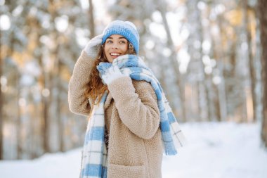 Portrait of a joyful woman with a bright scarf and hat enjoying herself in a snowy forest.  Beautiful woman enjoys a sunny winter day outdoors. Concept for relaxation and weekends.