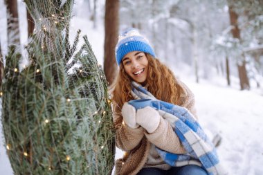 Young woman sits on log and drinks hot drink from thermos in snowy forest. A happy woman enjoys winter forest and drinks tea with a Christmas tree outdoors. Concept of celebration and relaxation.