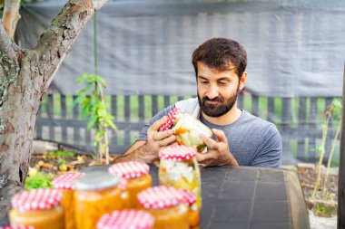 Sakallı mutlu Hintli Pakistanlı adam bahçesinden doğal ürünler üreten konserve, reçel, kavanoz ve limonlarla dolu bir masanın yakınındaki turşu kavanozunu inceliyor.