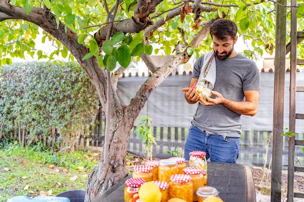 Happy eastern indian pakistani man with beard leisurely gazes and examines jar of pickles near table full of conserves, jams and jars and lemons, producing natural products from his garden under tree