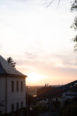 Çatısı ve penceresi olan bir ev batan güneşle çizilmiş renkli bulutlar ve gökyüzü, önünde ağaçlar, sakin bir alacakaranlık manzarası yaratıyor.