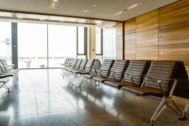 Büyük pencereler ve doğal ışık yakınında modern koltuklu boş bir havaalanı salonu. Huzurlu seyahat ortamı.