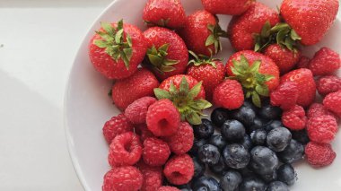Taze çilekler, ahududular ve yabanmersinleri doğal ışık altında beyaz bir kasede dizilmiştir. Görüntü sağlıklı yeme ve mevsimlik yaz üretimi kavramını aktarıyor..