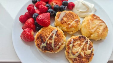 Taze çilek, ahududu, yabanmersini ve beyaz tabakta bir tutam kremayla servis edilen altın peynir krepleri. Görüntü, tatlı bir kahvaltı, hoşgörü ve ev yapımı rahatlığı temsil ediyor.