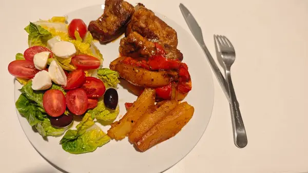 Kızarmış pirzola, fırında patates ve biberle birlikte domatesli, mozarellalı ve zeytinli taze bir salata. Görüntü ev yapımı ev yapımı yemekleri ve dengeli yemekleri temsil ediyor.