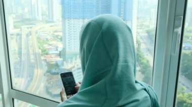 başörtüsü takmış genç Arap kadın pencere kenarında akıllı telefon kullanıyor.