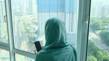 başörtüsü takmış genç Arap kadın pencere kenarında akıllı telefon kullanıyor.