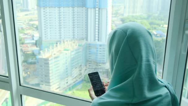 年轻的穆斯林妇女头戴头巾头巾 在家里靠窗的地方用智能手机 — 图库视频影像