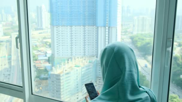 年轻的穆斯林妇女头戴头巾头巾 在家里靠窗的地方用智能手机 — 图库视频影像