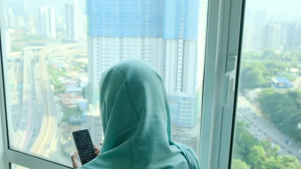 窓の近くの自宅でスマートフォンを使用してヘッドスカーフヒジャブの若いアラブイスラム教徒の女性 — ストック動画