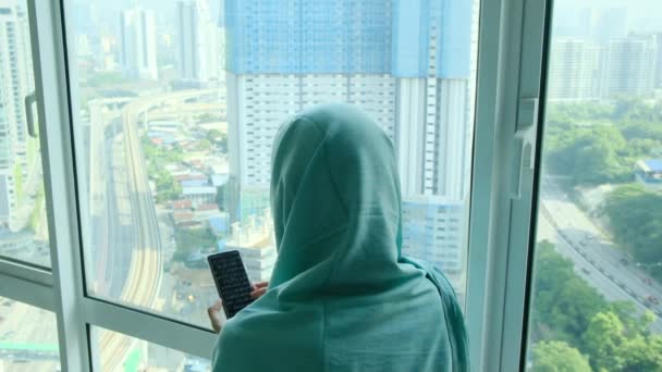 窓の近くの自宅でスマートフォンを使用してヘッドスカーフヒジャブの若いアラブイスラム教徒の女性 — ストック動画