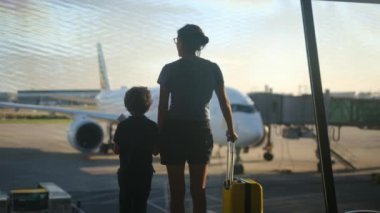 Anne ve oğlu havaalanı penceresinde bavuluyla bekliyor, uçaklara bakıyor.