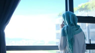tesettürlü çekici müslüman kadın pencerenin yanında cep telefonu kullanıyor..
