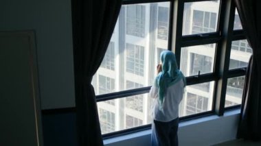 tesettürlü çekici müslüman kadın pencerenin yanında cep telefonu kullanıyor..