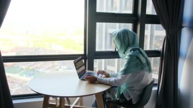 Pencerenin yanında dizüstü bilgisayar kullanan tesettürlü çekici Müslüman kadın..