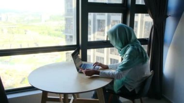 Pencerenin yanında dizüstü bilgisayar kullanan tesettürlü çekici Müslüman kadın..