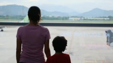 Anne ve oğlu havaalanı penceresinde bekliyor, uçaklara bakıyor.