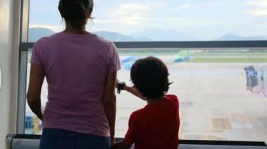 Anne ve oğlu havaalanı penceresinde bekliyor, uçaklara bakıyor.