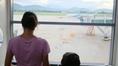 Anne ve oğlu havaalanı penceresinde bekliyor, uçaklara bakıyor.