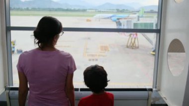 Anne ve oğlu havaalanı penceresinde bekliyor, uçaklara bakıyor.