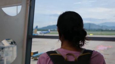 Solo gezgin havaalanı penceresinin yanında bavuluyla bekliyor, uçaklara bakıyor.