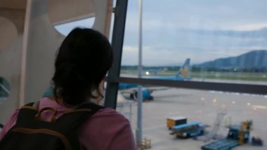 Solo gezgin havaalanı penceresinin yanında bavuluyla bekliyor, uçaklara bakıyor.
