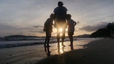 Anne ve oğulları güneşli bir günde deniz kıyısında, gün batımının altında okyanus sahillerinde yürüyorlar..