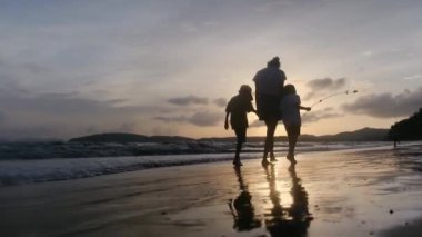 Anne ve oğulları güneşli bir günde deniz kıyısında, gün batımının altında okyanus sahillerinde yürüyorlar..