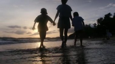 Anne ve oğulları güneşli bir günde deniz kıyısında, gün batımının altında okyanus sahillerinde yürüyorlar..