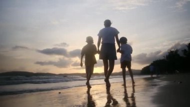 Anne ve oğulları güneşli bir günde deniz kıyısında, gün batımının altında okyanus sahillerinde yürüyorlar..