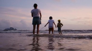 Anne ve oğulları güneşli bir günde deniz kıyısında, gün batımının altında okyanus sahillerinde yürüyorlar..