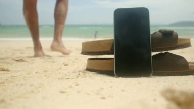 Parmak arası terlik ve cep telefonu, kumlu yaz plajı arka planında. Mutlu tatiller. Yakın çekim.