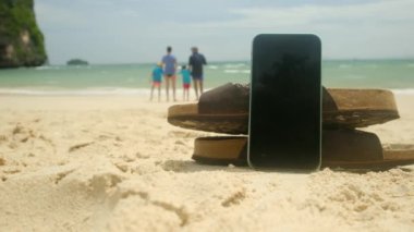 Parmak arası terlik ve cep telefonu, kumlu yaz plajı arka planında. Mutlu tatiller. Yakın çekim.