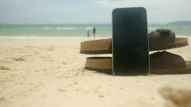 砂浜の背景にフリップフロップと携帯電話 ハッピー 楽しい休暇 閉じる — ストック動画