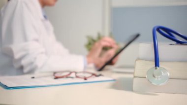 Masadaki kitaplarla steteskopu kapat ve kadın doktor hastanedeki arka planda tablet bilgisayar kullanıyor