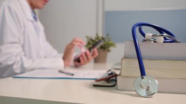 Masadaki kitaplarla steteskopu kapat ve kadın doktor hastanede arka planda akıllı telefon kullanıyor.