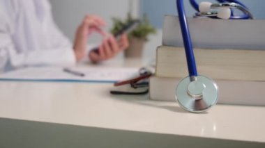 Masadaki kitaplarla steteskopu kapat ve kadın doktor hastanede arka planda akıllı telefon kullanıyor.