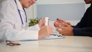 Beyaz üniformalı genç kadın doktoru yakından görüyor ve sağlık kliniğinde masasında oturan erkek hastayla sonuçları ya da belirtileri tartışıyor.