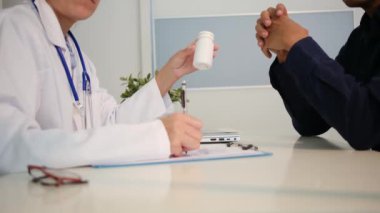 Beyaz üniformalı genç kadın doktoru yakından görüyor ve sağlık kliniğinde masasında oturan erkek hastayla sonuçları ya da belirtileri tartışıyor.