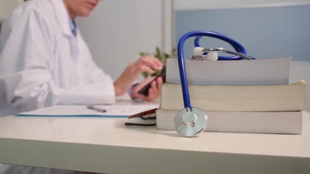 近景听诊器与书籍放在桌上 女医生在医院背景下使用智能手机 — 图库视频影像
