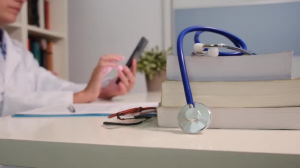近景听诊器与书籍放在桌上 女医生在医院背景下使用智能手机 — 图库视频影像