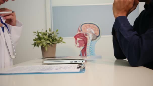 白い医療ユニフォームで若い女性医師の視界を閉じ 健康診療所の机に座っている男性患者と結果または症状を議論する — ストック動画