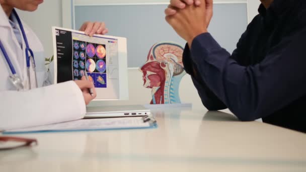白い医療ユニフォームで若い女性医師の視界を閉じ 健康診療所の机に座っている男性患者と結果または症状を議論する — ストック動画