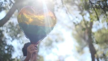 Kişi tarafından tutulan LGBTQ + gurur bayrağı renginde kalp şeklinde bir balon. Güneş ışınları eşitliği, cinsel özgürlüğü, cinsiyet saygısını ve dahil edilmeyi simgeliyor.